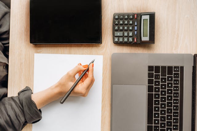 a-person-holding-a-pencil-ready-to-write-on-a-blank-sheet-of-paper-with-a-laptop-calculator-and-tablet-on-the-desk