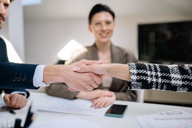 two-people-shaking-hands-over-a-desk-with-documents-while-a-smiling-woman-observes-in-the-background