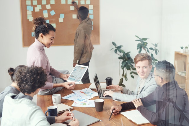 a-group-of-colleagues-collaborating-at-a-table-with-a-person-writing-on-a-wall-of-sticky-notes-in-the-background