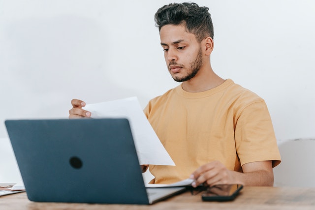 a-man-in-a-tan-shirt-sitting-at-a-desk-reviewing-a-document-while-working-on-a-laptop-with-a-smartphone-beside-him