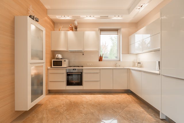a-bright-kitchen-with-glossy-white-cabinets-beige-walls-and-floors-a-built-in-oven-and-a-window-above-the-sink-letting-in-natural-light