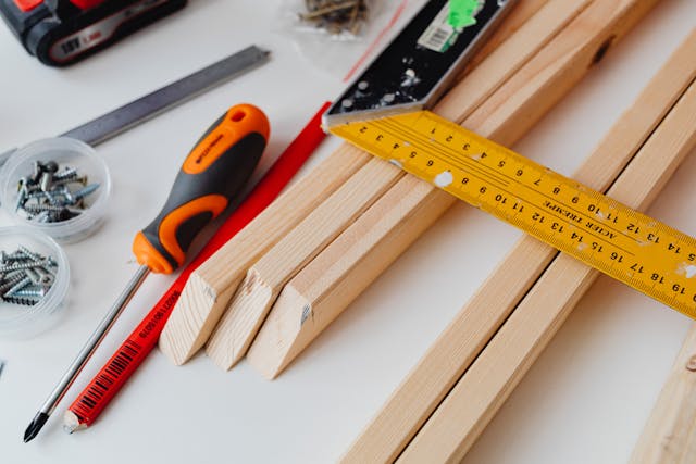 a-close-up-of-woodworking-tools-and-materials-on-a-white-surface-including-wooden-planks-screws-a-screwdriver-a-pencil-a-measuring-square-and-a-ruler