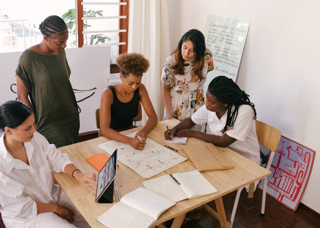 a-group-of-five-women-gathered-around-a-wooden-table-collaborating-on-sketches-and-notes-with-a-laptop-and-notebooks-open-in-a-well-lit-room