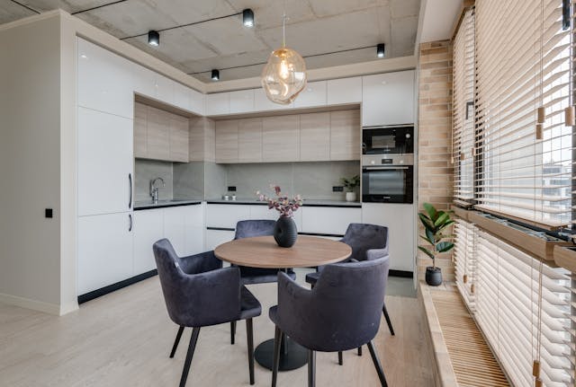 a-modern-kitchen-and-dining-area-with-a-round-table-four-velvet-chairs-sleek-white-cabinets-and-large-windows-with-wooden-blinds