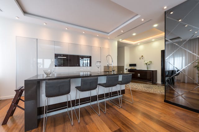 a-modern-kitchen-with-a-large-island-six-black-bar-stools-glossy-white-cabinetry-wood-flooring-and-recessed-ceiling-lights
