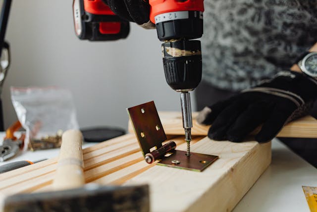 a-person-using-a-power-drill-to-attach-a-metal-hinge-to-wooden-boards-with-tools-and-screws-visible-on-the-work-surface