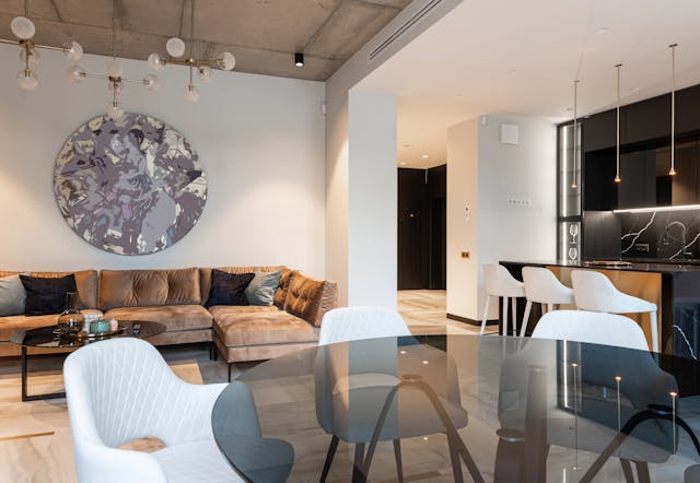 a-stylish-open-concept-living-space-with-a-glass-dining-table-a-velvet-sectional-sofa-modern-pendant-lights-and-a-dark-kitchen-with-a-marble-backsplash
