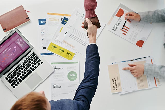 business-meeting-scene-with-two-people-shaking-hands-over-a-desk-covered-in-financial-reports-charts-and-a-laptop