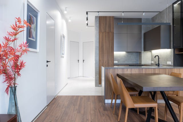 modern-open-concept-kitchen-and-dining-area-with-wood-and-gray-cabinetry-a-dark-dining-table-with-tan-chairs-and-decorative-red-orange-flowers-in-a-vase