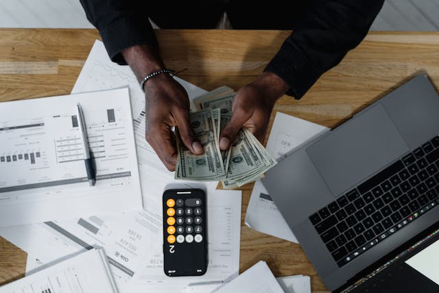 person-sitting-at-desk-counting-money-and-using-a-calculator-with-papers-and-a-laptop-around-them