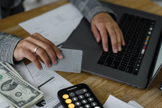 person-using-a-laptop-while-holding-a-bank-card-with-cash-documents-and-a-calculator-on-the-table