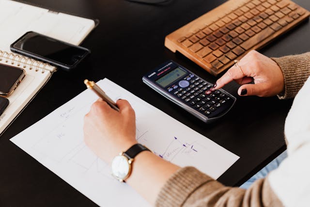 person-using-a-scientific-calculator-and-writing-on-a-sheet-of-paper-at-a-desk-with-a-keyboard-notebook-and-smartphones-nearby