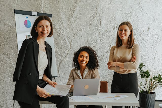three-professional-women-smiling-in-a-modern-office-setting-with-one-seated-at-a-desk-with-a-laptop-and-the-others-standing-nearby-next-to-a-flip-chart-and-some-documents