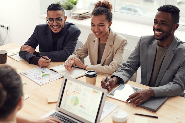 three-professionally-dressed-people-sit-at-a-table-smiling-during-a-meeting-with-charts-notebooks-and-coffee-cups-in-front-of-them-and-a-laptop-screen-visible-in-the-foreground