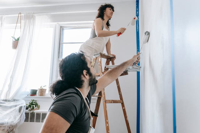 two-people-painting-a-wall-white-one-standing-on-a-ladder-and-the-other-reaching-up-with-a-roller-in-a-bright-room-with-natural-light