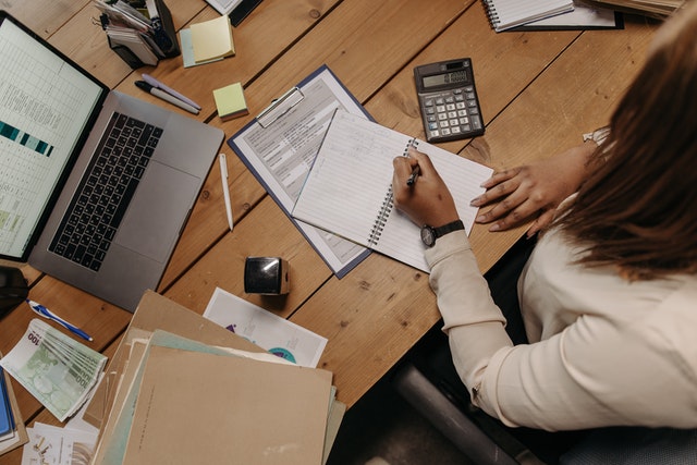 a-person-writing-notes-in-a-notebook-on-a-wooden-desk-surrounded-by-a-laptop-calculator-documents-and-office-supplies
