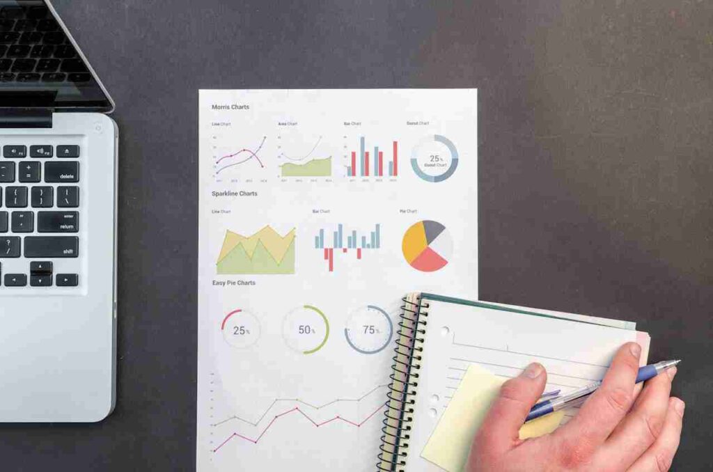 laptop-financial-charts-and-a-person-writing-in-a-notebook-on-a-desk