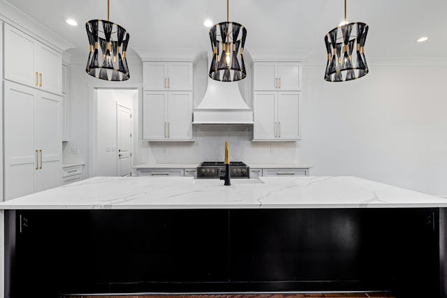 a-modern-kitchen-with-a-white-marble-island-three-pendant-lights-with-black-and-gold-shades-white-cabinets-and-a-stainless-steel-stove