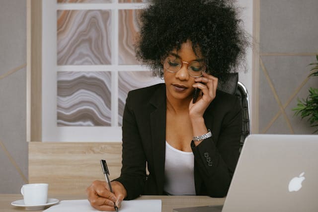 a-person-in-a-black-blazer-talking-on-the-phone-while-taking-notes-at-a-desk-with-a-laptop-and-coffee-cup