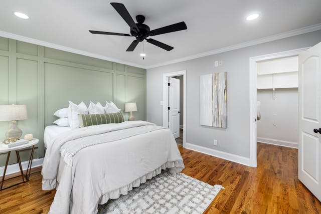 bedroom-with-a-white-bed-green-accent-wall-nightstands-with-lamps-a-ceiling-fan-and-wooden-flooring-there-is-an-open-door-leading-to-a-walk-in-closet-and-a-door-to-another-room