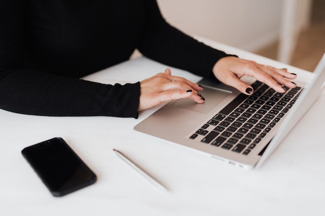 person in a black shirt working on their laptop with their phone next to them on a white desk