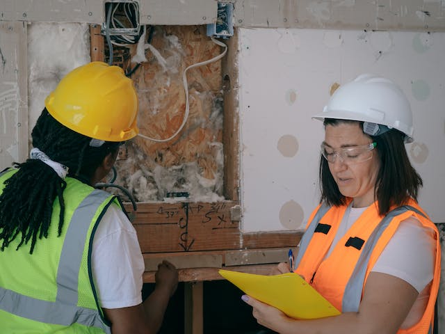 two-people-in-hard-hats-and-safety-vests-inspecting the-wiring-and-insulation-of-a-wall