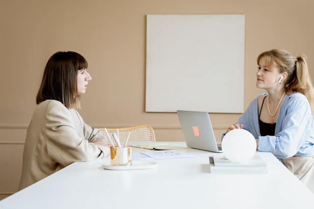 two-people-sitting-at-a-white-table-having-a-work-conversation-with-laptops-pens-and-notebooks