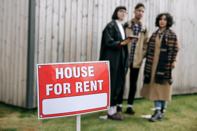 a red house for rent sign on a green lawn with prospects standing in the background