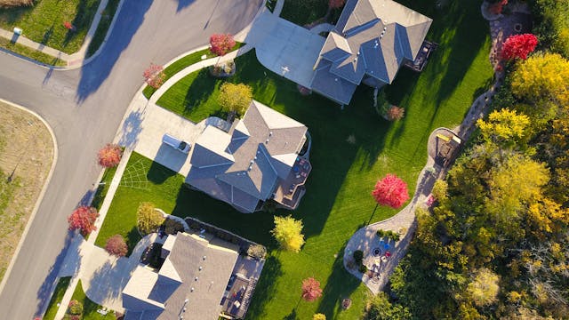 Aerial view of suburban houses with green lawns, trees with autumn colors, and a nearby wooded area.