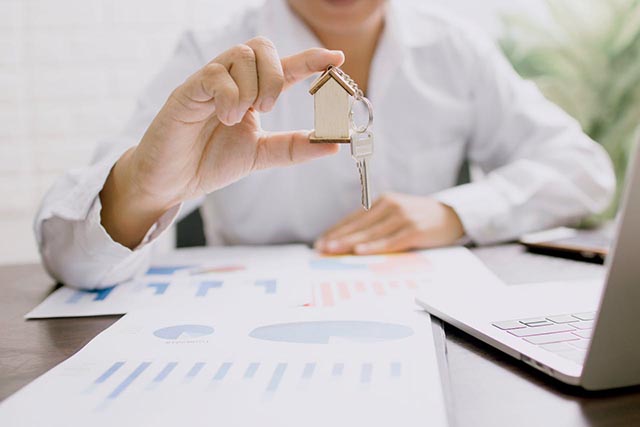 a-person-holding-a-small-wooden-house-keychain-with-a-key-over-a-desk-with-charts-and-a-laptop