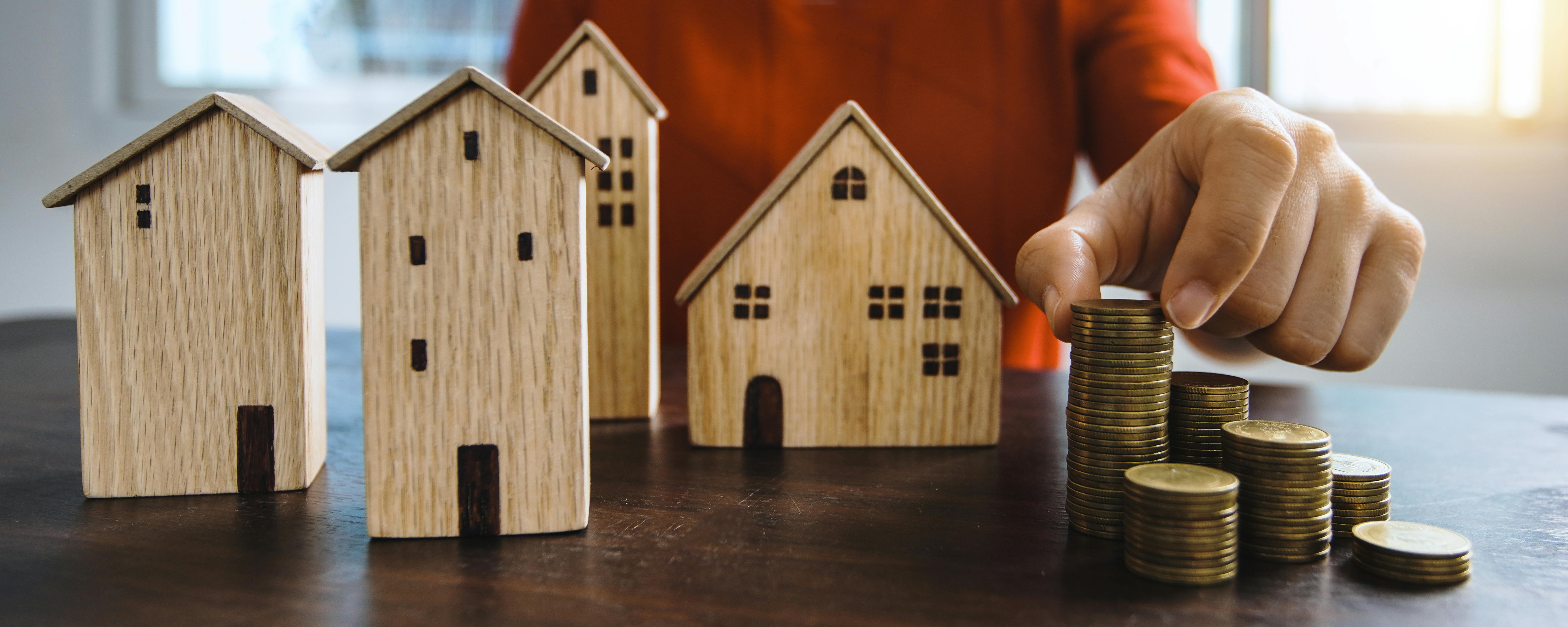A person stacking gold coins beside four miniature wooden houses.